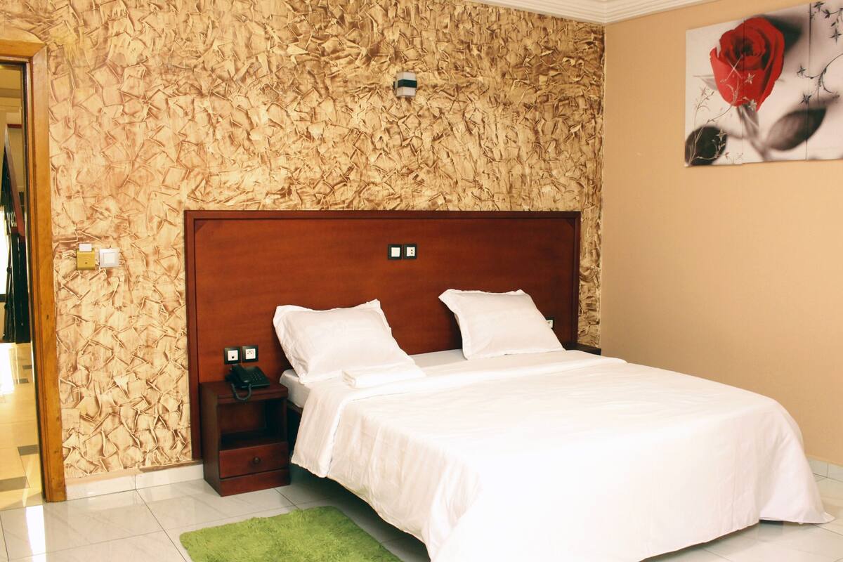 A neatly made bed with white linens, a wooden headboard, a green rug, and a textured wall behind it.