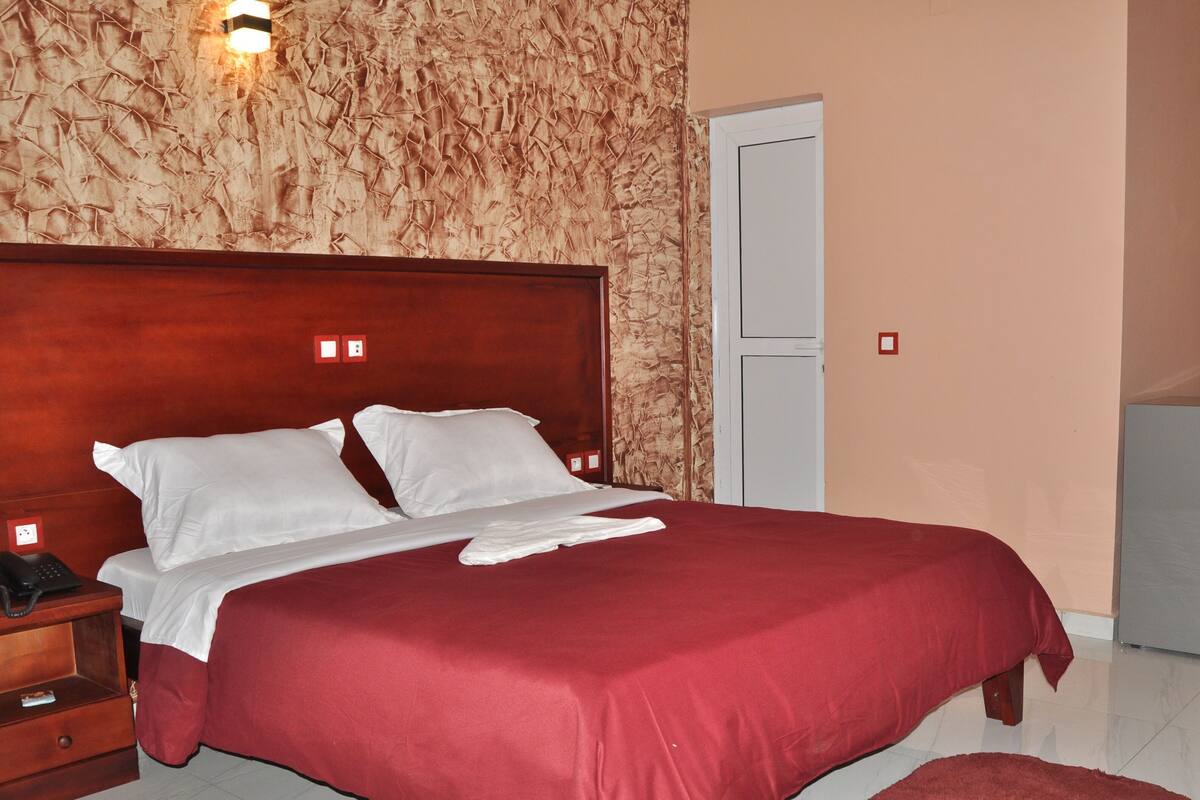 A hotel room with a large bed, a wooden nightstand, a desk, a chair, a refrigerator, and a door.