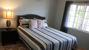 A neatly made bed with striped bedding, a bedside lamp, and a window with curtains. - Soleil de la Palms (St. Ann's Bay)