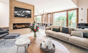 A modern living room with a fireplace, sofa, TV, and dining area with large windows.