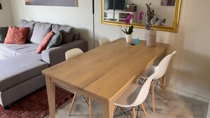 Una sala de estar moderna con un sofá gris, una mesa de comedor de madera y sillas blancas. Hay un espejo grande en la pared, un televisor y una planta en maceta.