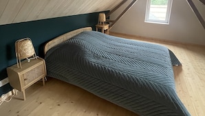 A loft bedroom with a sloped ceiling, a bed with a quilted blue cover, a wicker chair, a small table, and a window providing natural light. - Gîte Les Roches D'Étigues near Etretat (Vattetot-sur-Mer)