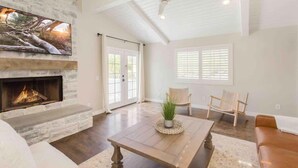 A spacious living room with a fireplace, a ceiling fan, and a large window with blinds. - Hakuna Moscato - 30 day Wine Country Retreat (Temecula)