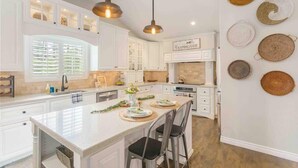 A spacious kitchen with white cabinetry, a large island, pendant lighting, and woven placemats on the wall. - Hakuna Moscato - 30 day Wine Country Retreat (Temecula)