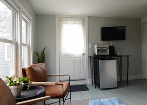 Ein gemütlicher Wohnraum mit einer Couch, einem Sessel, einem kleinen Tisch mit Pflanze, einem Kühlschrank, einer Mikrowelle und einem Fernseher auf einem Schrank.