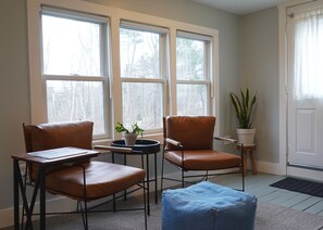 Ein Zimmer mit zwei Ledersesseln, einem kleinen Tisch und einem blauen Hocker. Es gibt drei Fenster mit Jalousien, einen Blumentopf auf dem Fensterbrett und eine Tür mit Vorhang.