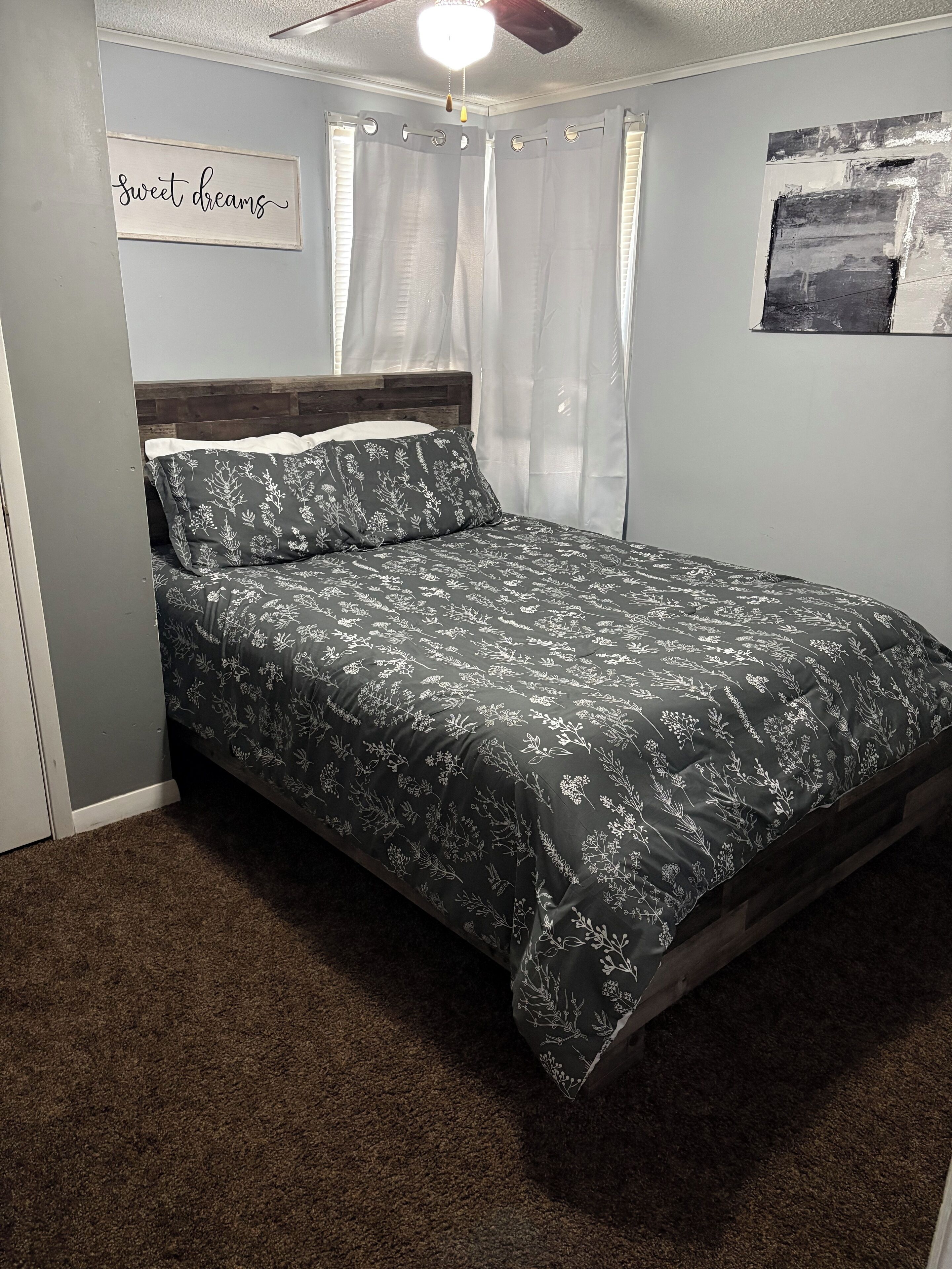 A bedroom with a bed, a ceiling fan, a wall-mounted sign, and a window with curtains.