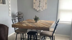 Un coin repas comprenant une table en bois, quatre chaises, un ventilateur de plafond, une horloge murale et une fenêtre avec des stores.