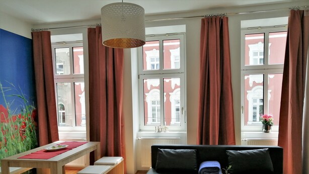 Ein modernes Wohnzimmer mit einer Couch, einem Tisch und einer Bank. Große Fenster mit Vorhängen, eine Heizung und eine Wand mit floralem Wandbild.