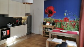 Eine moderne Küche mit weißen Schränken, einer schwarzen Arbeitsplatte und einem Edelstahl-Kühlschrank. Ein Holztisch mit einer roten Tischmatte und einem weißen Laptop sind vorhanden. An der Wand hängt ein großes Gemälde einer Mohnblume.