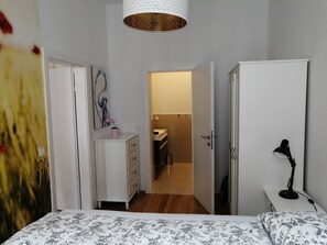 Um quarto com cama, cômoda, escrivaninha com luminária e banheiro visível por uma porta aberta.