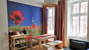 Ein Zimmer mit Holzboden, einem Tisch mit roter Unterlage, einem schwarzen Sofa, einem Wandbild mit Mohnblumen und einem Fenster mit Vorhängen.