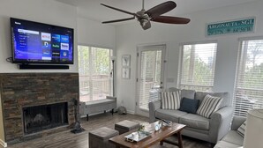 Een woonkamer met een stenen open haard, een grijze bank, een houten salontafel en een flatscreen televisie aan de muur.