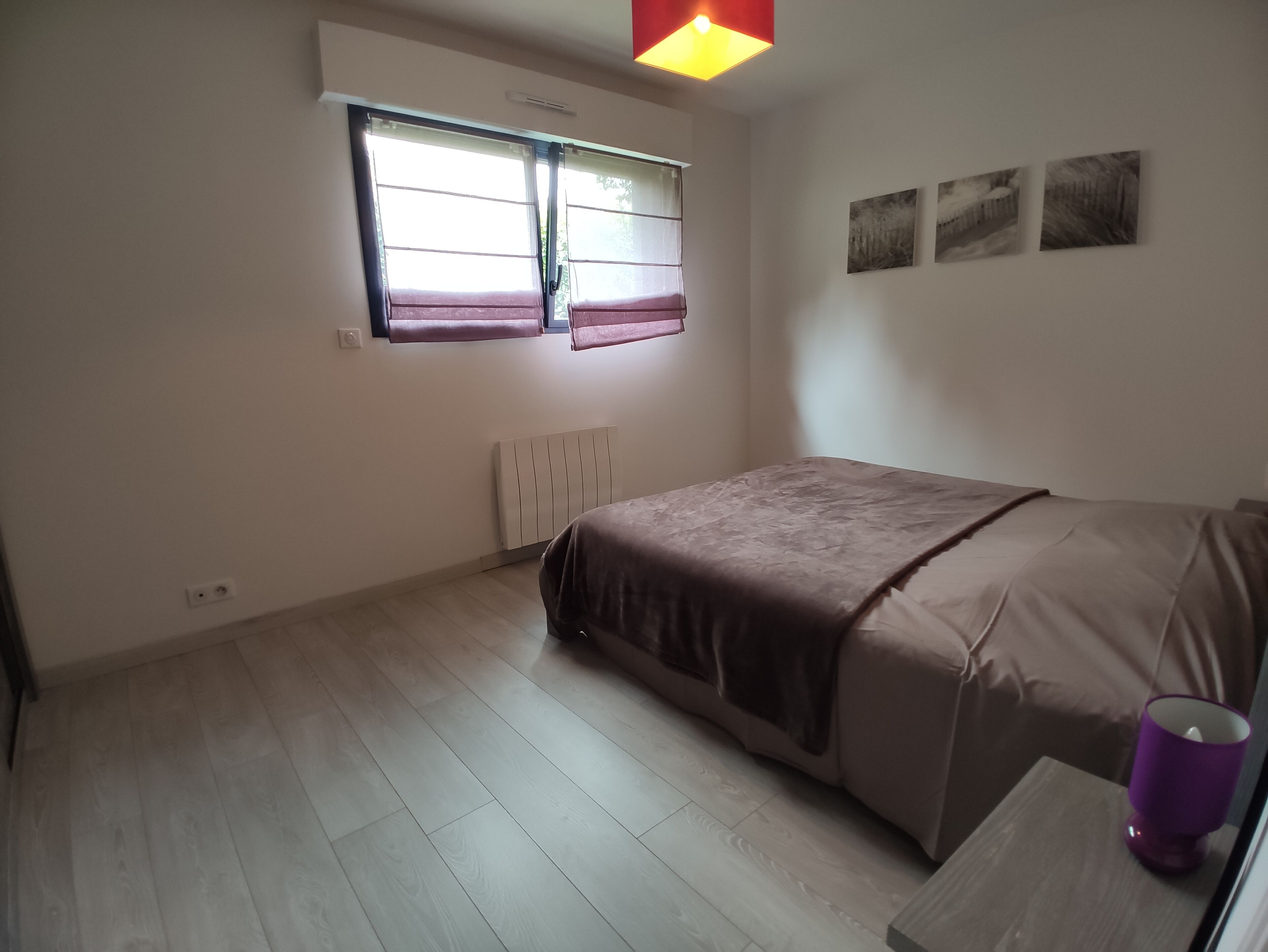 A bedroom with a bed, a window with curtains, a radiator, and a wall with three pictures.