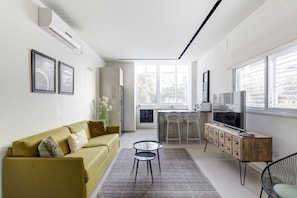 A modern living room with a sofa, a small table, a TV, and a kitchen area with bar stools.