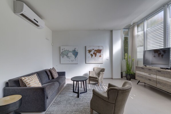 A modern living room with a grey sofa, a round coffee table, a beige armchair, a TV on a wooden cabinet, and two world maps on the wall.
