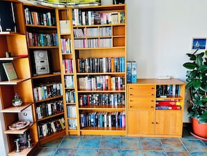 A room with a large wooden bookshelf filled with books, a small cabinet with books, a potted plant, and a wall-mounted picture. - Bright, spacious vacation apartment in Schwedeneck/Surendorf, 1 km to the beach (Schwedeneck)