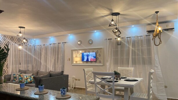 A modern living room with a dining area, featuring a TV, decorative lighting, and a large plant. - Caribbean Golf (Punta Cana)