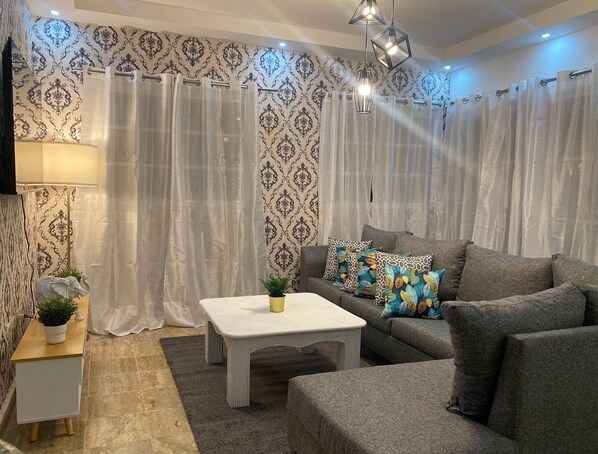 A living room with patterned wallpaper, a grey sofa with colorful cushions, a white coffee table, and a small side table with a plant.