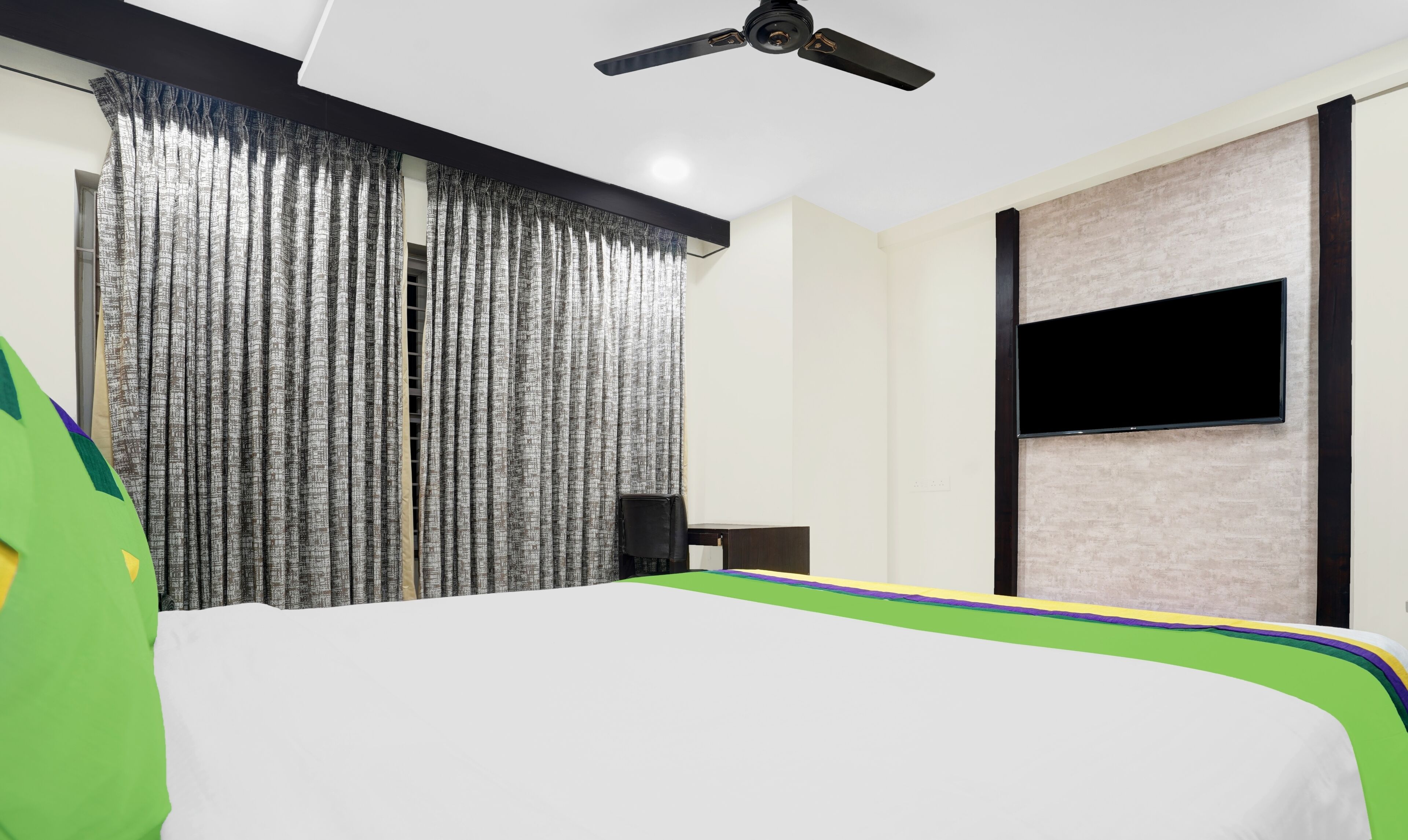Habitación de hotel con cama, televisor montado en la pared, ventilador de techo y ventana con cortinas.