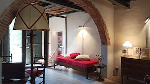 A bedroom with a red bed, a wooden chair, a desk with a lamp, and a brick archway.