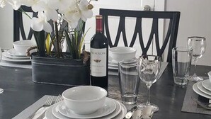 Ein Esstisch ist gedeckt mit weißen Tellern, Schüsseln, Besteck und Weingläsern. Eine Flasche Franciscan Wein ist sichtbar, ebenso eine Orchidee im Blumentopf.