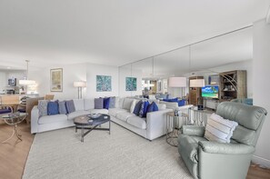 A modern living room with a white sofa, a leather armchair, and a coffee table. There are multiple lamps and a flat-screen TV. - Beachfront Condo | Pool, Gym, & Pickleball (Marco Island)