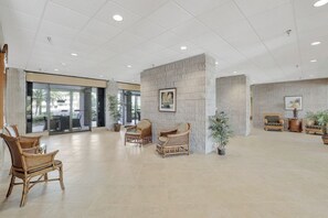 A spacious lobby with wicker chairs, a central column with a framed picture, and potted plants. - Beachfront Condo | Pool, Gym, & Pickleball (Marco Island)