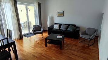 Ein Wohnzimmer mit Holzboden, einer schwarzen Couch mit Kissen, einem schwarzen Couchtisch und einem grauen Sessel.