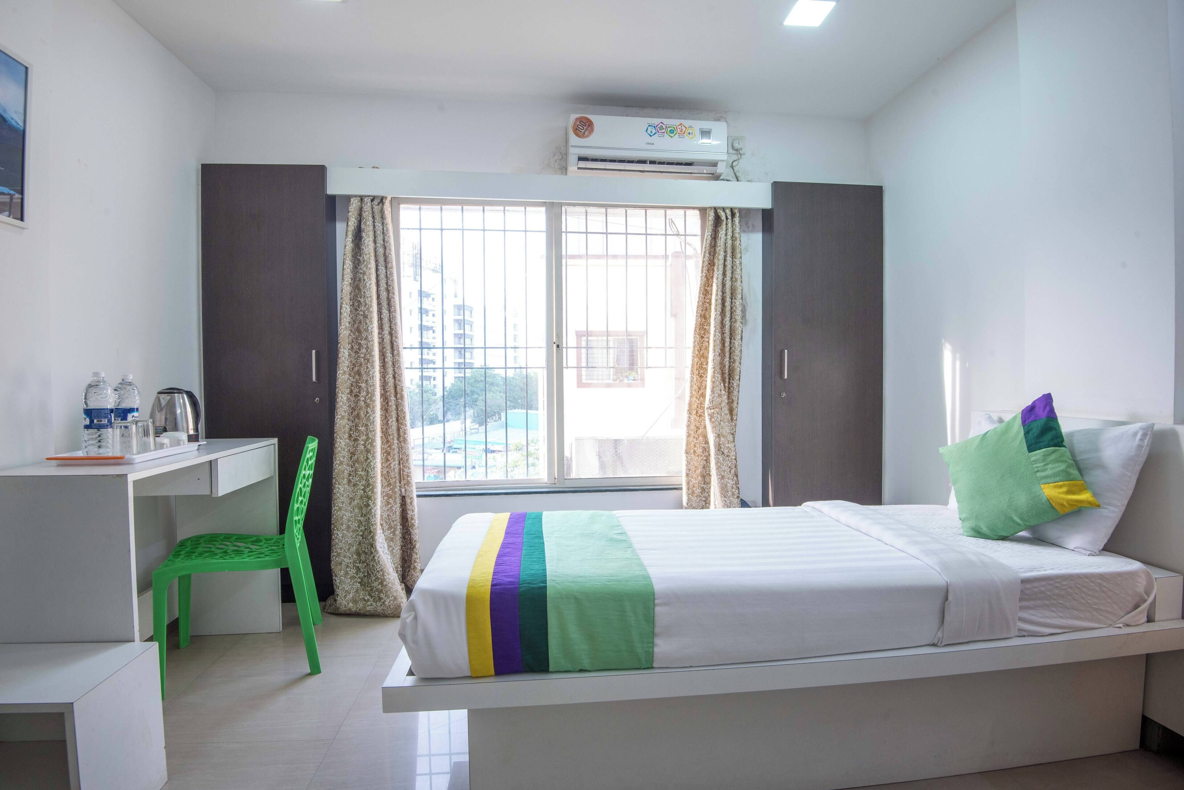 A hotel room with a bed, a desk, a chair, an air conditioning unit, and a window with curtains.