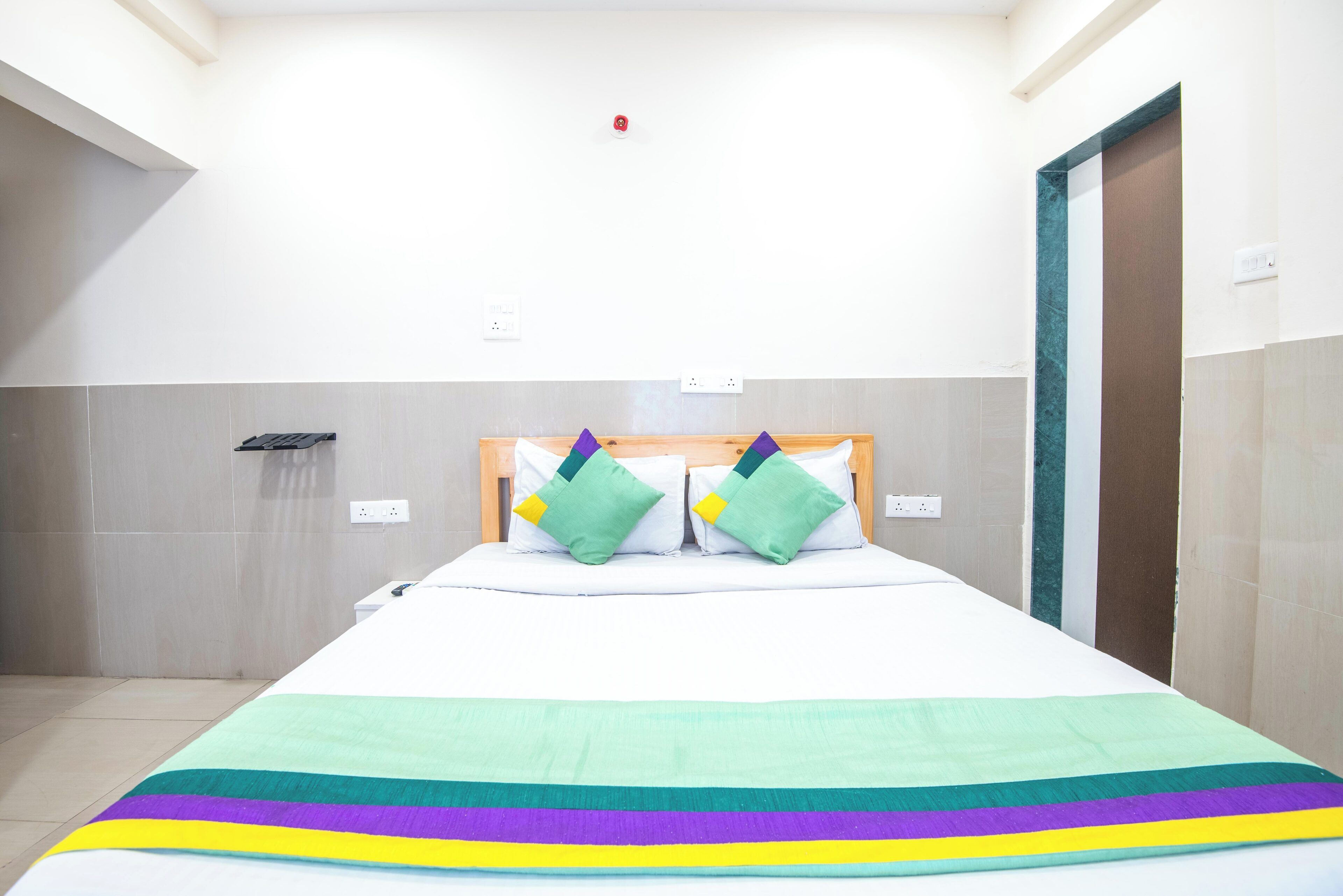 A hotel room with a bed, a wooden headboard, two green pillows, a white bedspread, and a colorful blanket.