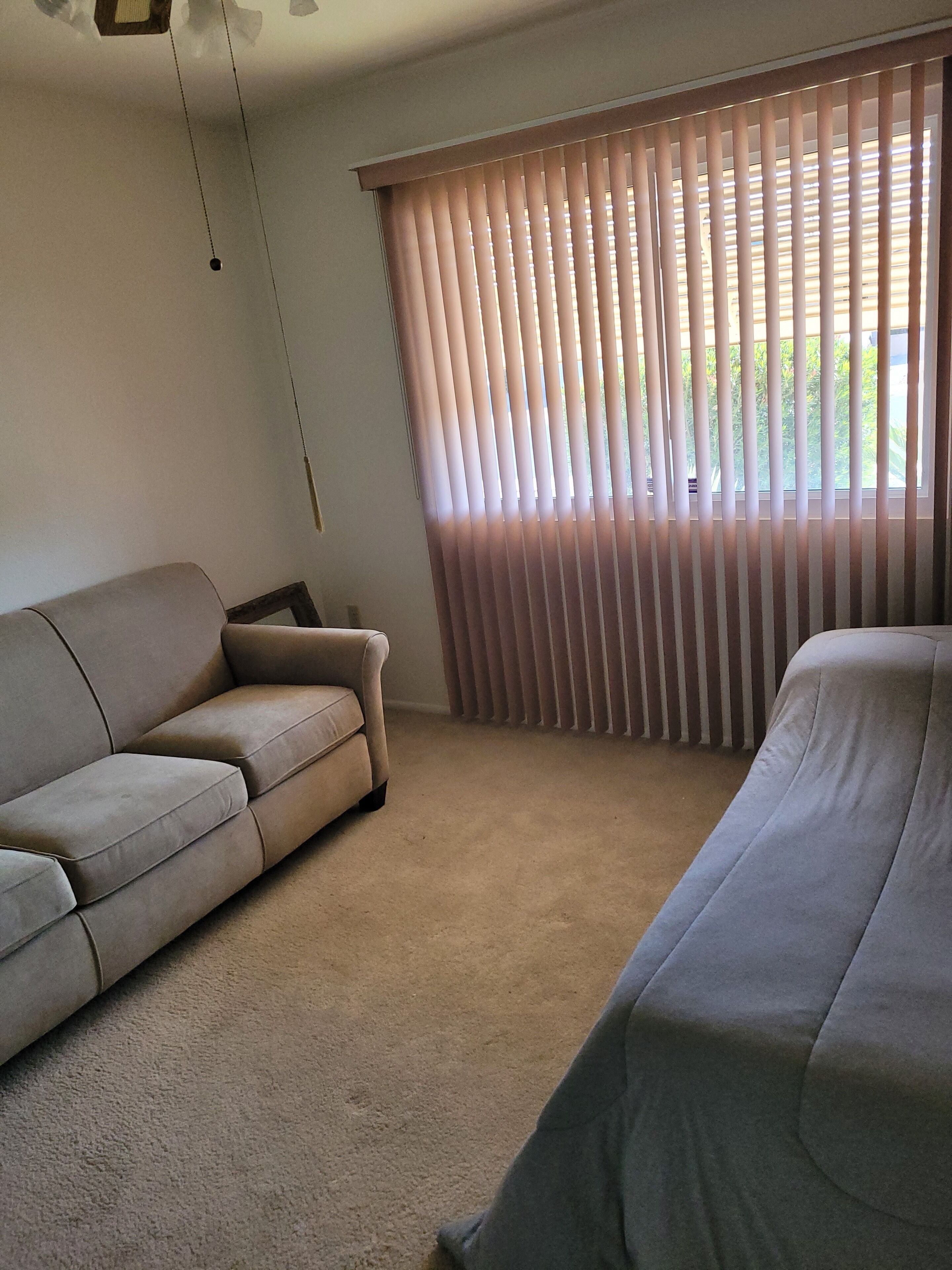 Une pièce avec un tapis beige, un canapé beige, un lit recouvert d’une couverture grise et des stores verticaux à la fenêtre.