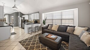 A modern living room with a sectional sofa, ottoman, and a kitchen in the background. - Texas Oasis With Pool! (Fort Worth)