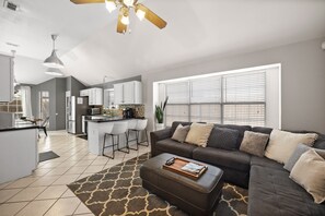 A modern living room with a sectional sofa, ottoman, and a kitchen in the background.