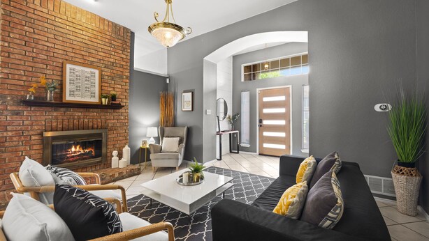 A modern living room with a brick fireplace, a black sofa with patterned cushions, and a white coffee table. - Texas Oasis With Pool! (Fort Worth)