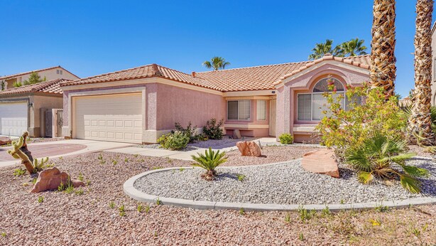 Une maison d’habitation avec un garage, une terrasse agrémentée d’un palmier et un jardin aménagé avec des rochers et des cactus.