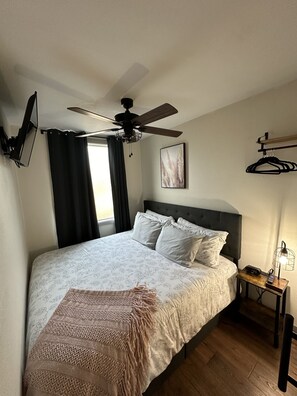 A bedroom with a bed, a ceiling fan, a nightstand with a lamp, and a window with curtains.