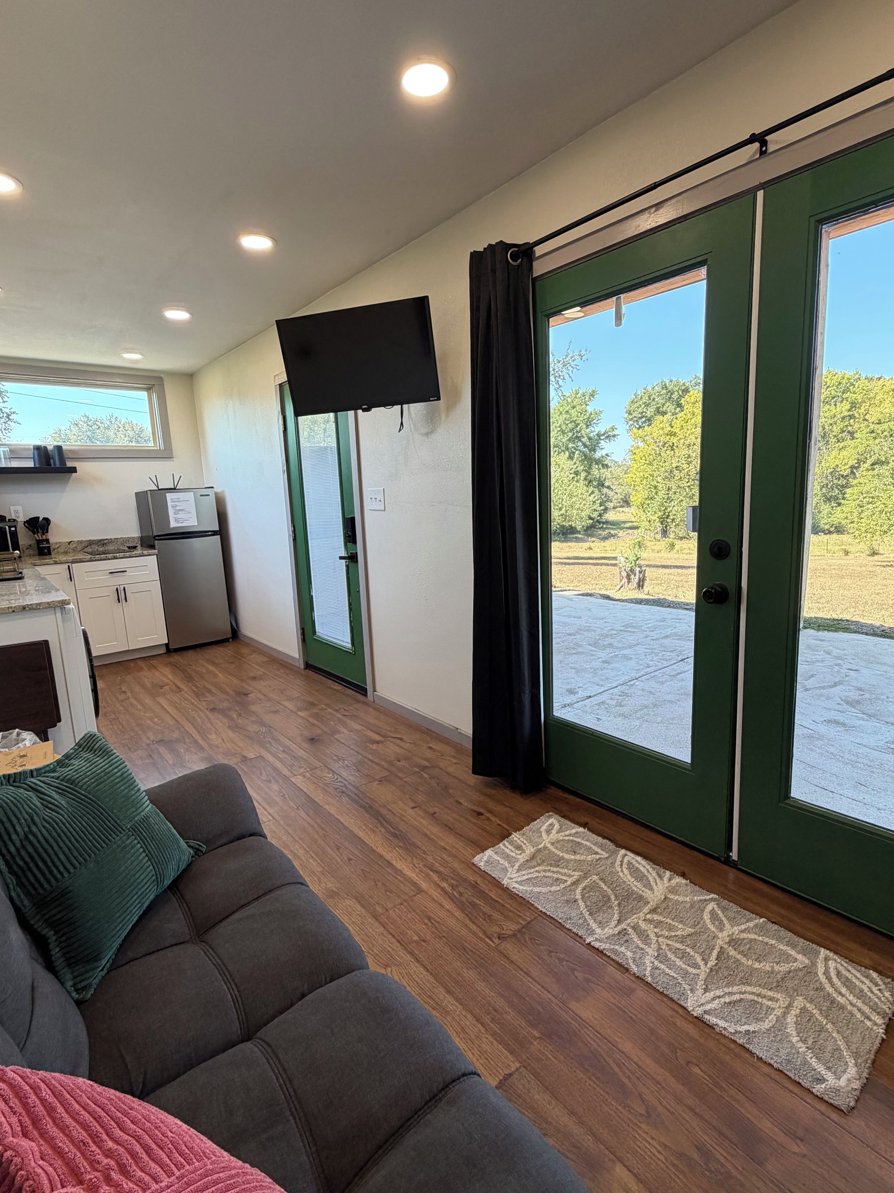A modern living space with a sofa, a flat-screen TV, a kitchen area, and a sliding glass door leading to an outdoor area.