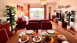 Ein modernes Wohnzimmer mit Sofa, Esstisch und Fernseher. Pflanzen, ein Balkon mit Aussicht und ein Essbereich mit Frühstücksangeboten.