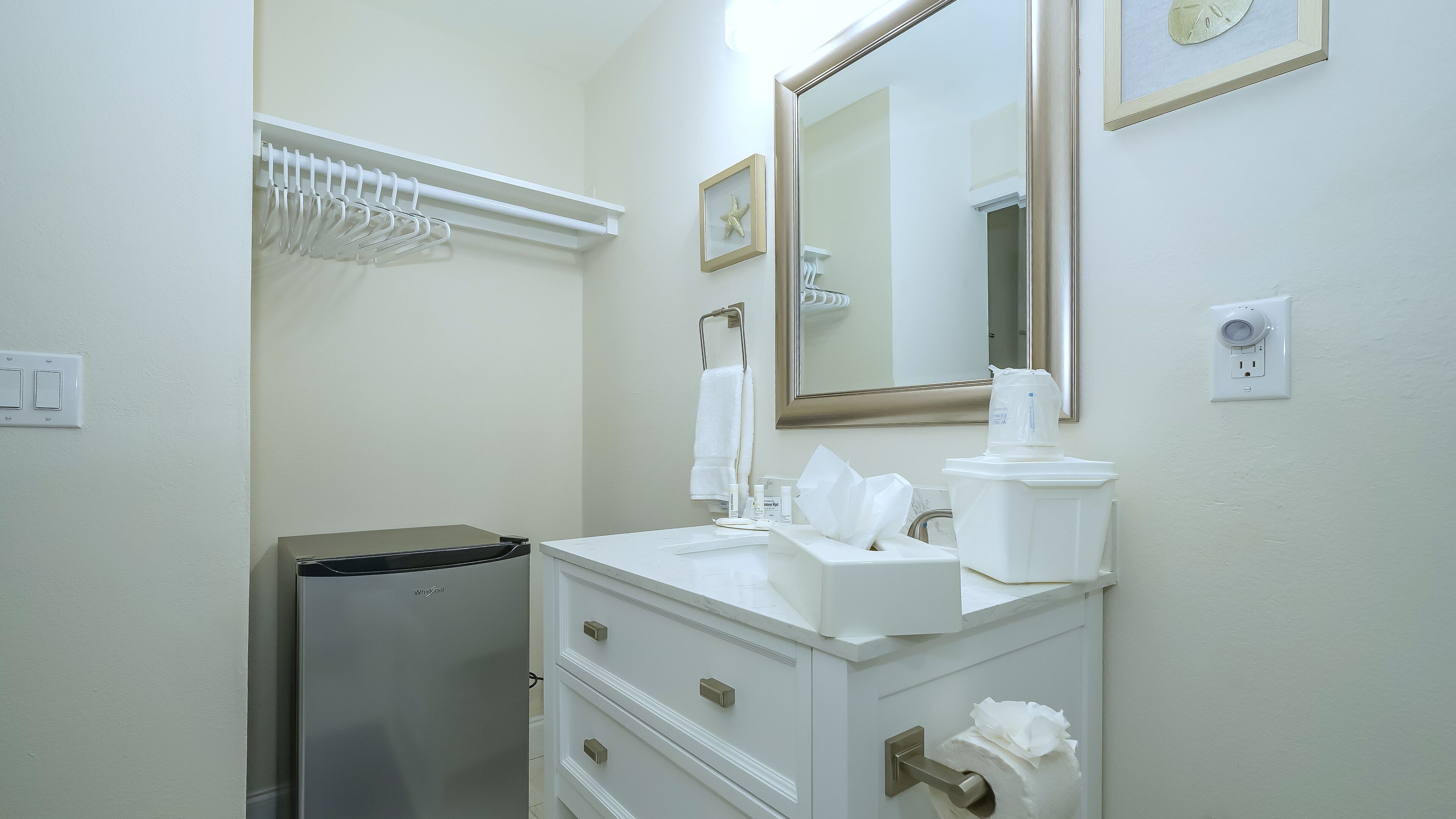 Un baño blanco con lavamanos, espejo, toallero y un refrigerador pequeño.