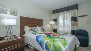 Habitación de hotel con una cama, un escritorio con lámpara, una silla y un ventanal con cortinas.