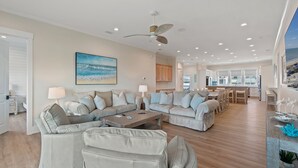 A spacious living room with a large sectional sofa, a coffee table, and a ceiling fan. There is a kitchen area visible in the background.