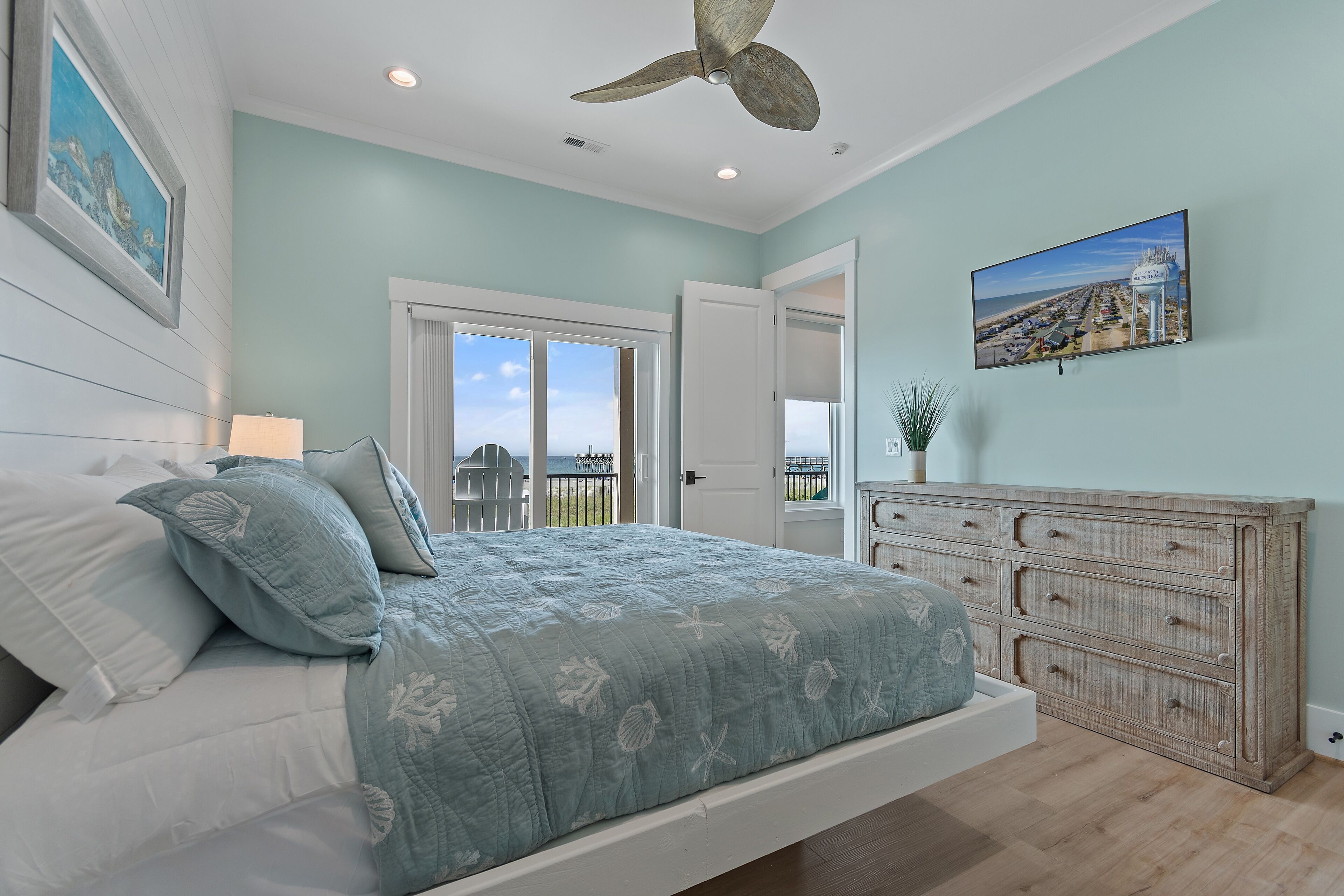 A bedroom with a bed, a dresser, a ceiling fan, a painting, and a view through a door.