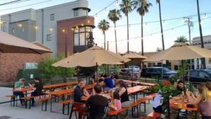 An outdoor dining area with tables and umbrellas, surrounded by palm trees and buildings.