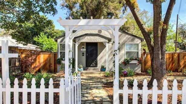 A house with a stone pathway leading to a black door, surrounded by a white picket fence and a pergola.