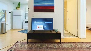 A living room with a flat-screen TV on a stand, a painting on the wall, and a kitchen area in the background.