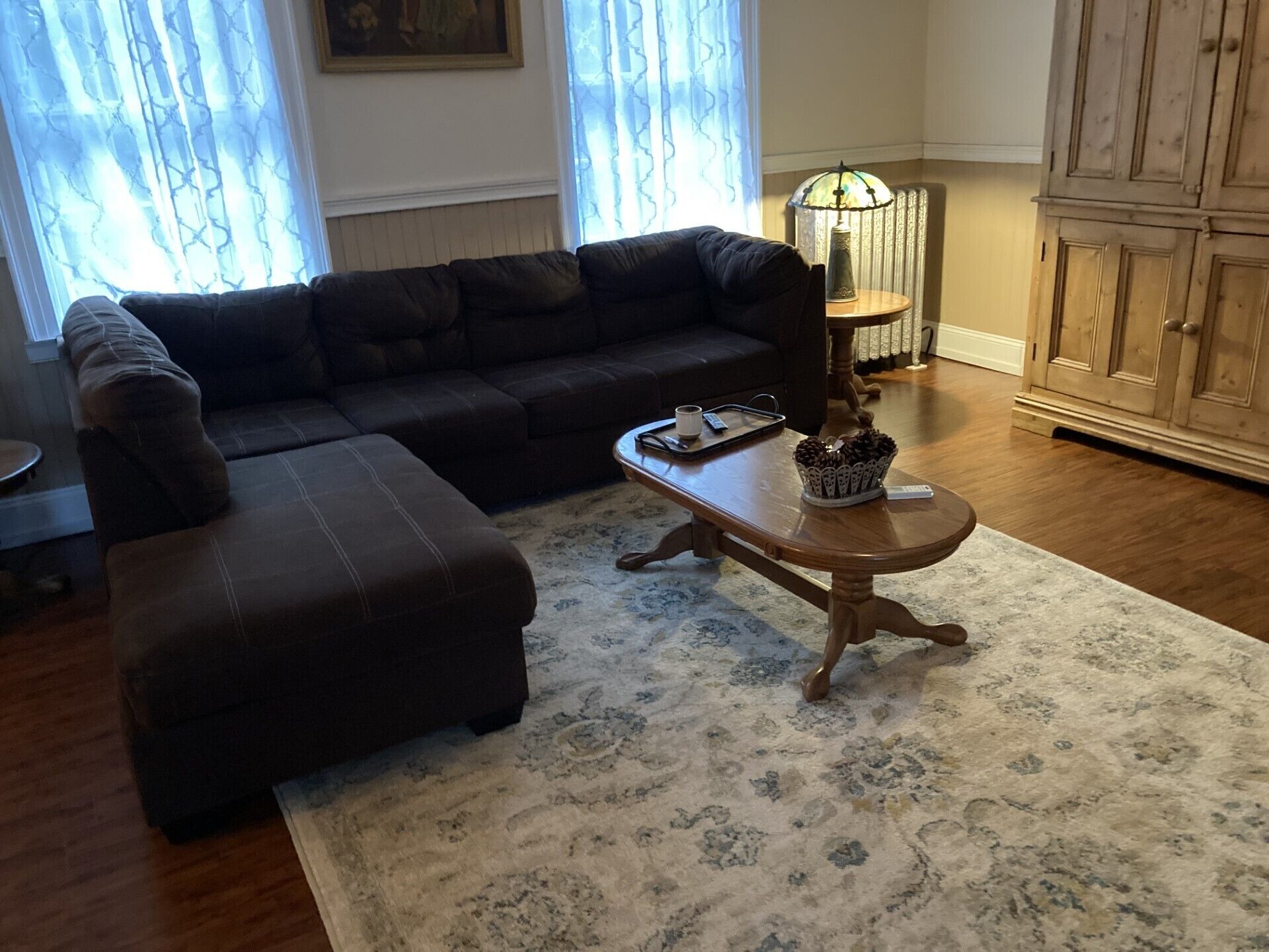 Una sala de estar con un sofá marrón oscuro, un reposapiés a juego, una mesa de centro de madera con una bandeja, una lámpara y una alfombra estampada.