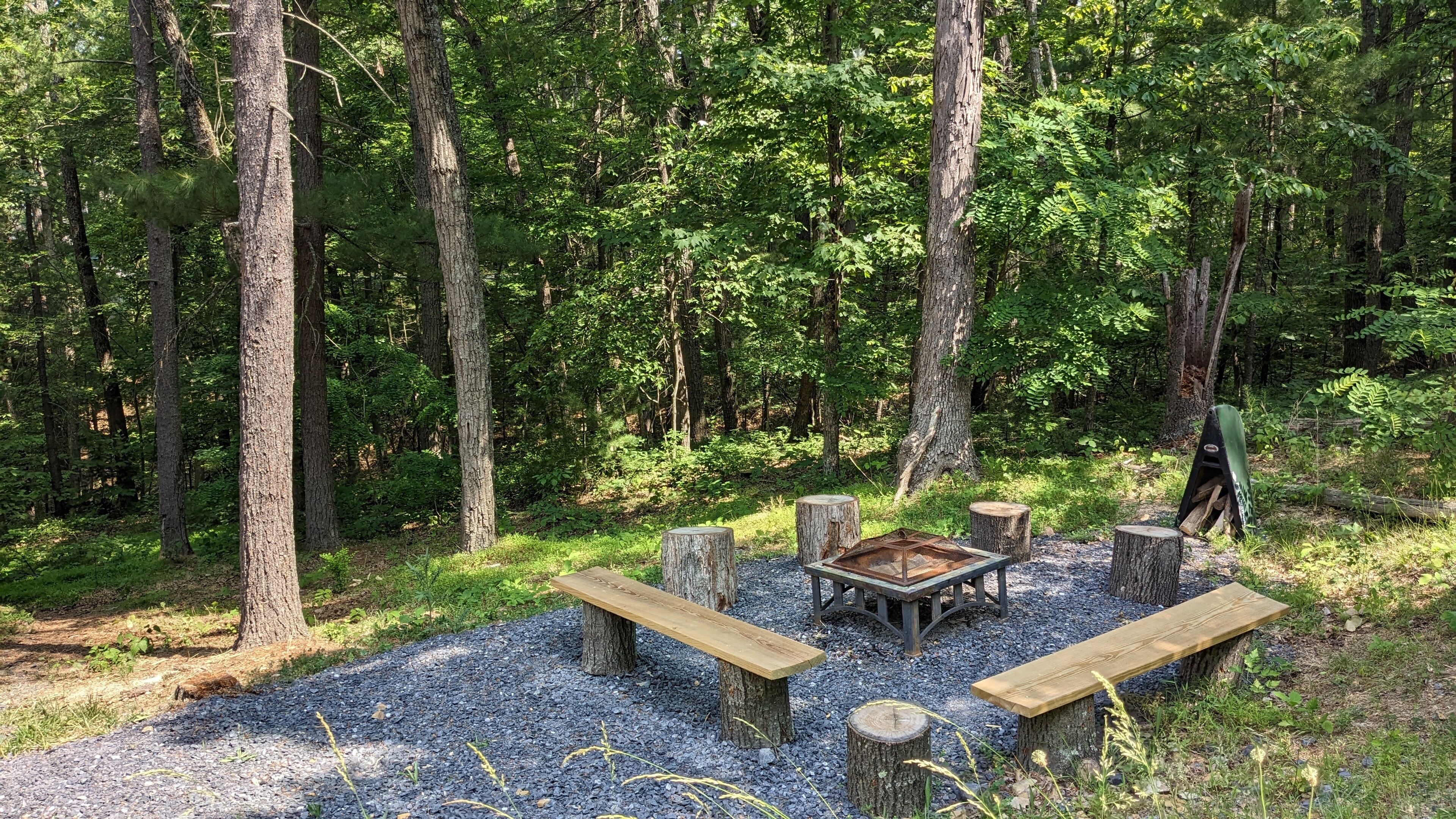 Een kampeerterrein omringd door bos, met een metalen vuurkuil, houten banken en een picknicktafel.