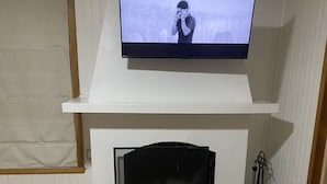 Un televisor montado en una pared blanca sobre una chimenea, una mesa de madera al frente y un control remoto sobre la mesa.