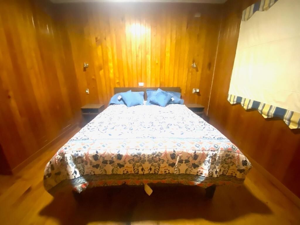 A bedroom with a wooden floor and walls, a bed with floral bedding and blue pillows, and a window with a striped curtain.